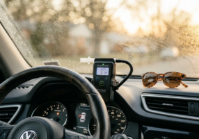 Close-up of a car dashboard area at eye level, a small breath-test device mounted near the steering column, morning condensation on the windshield, realistic vehicle interior with personal items like sunglasses on the dash
